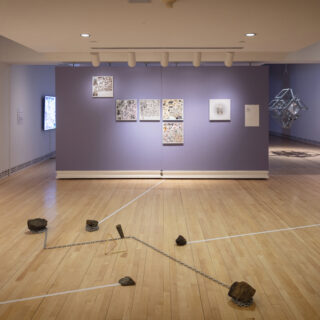 A gallery space with a collection of square artworks hung on a purple wall, and an installation comprised of rocks, chains, and a small abstract sculpture in the foreground.