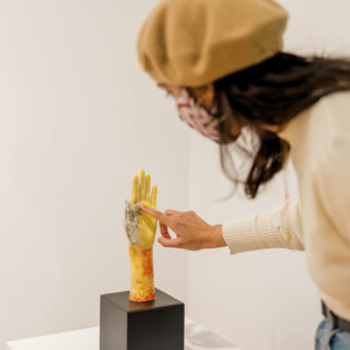 A person with a beige beret and sweater touches a yellow, orange, and grey hand sculpture with intricate painted details, placed on a black stand in an art gallery with white walls.