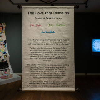 Installation view of a panel titled "The Love that Remains" in the middle, featuring curatorial statements with colorful embroidery. To each side are a knitted fabric piece and a blue illuminated artwork.