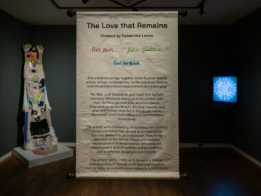 Installation view of a panel titled "The Love that Remains" in the middle, featuring curatorial statements with colorful embroidery. To each side are a knitted fabric piece and a blue illuminated artwork.