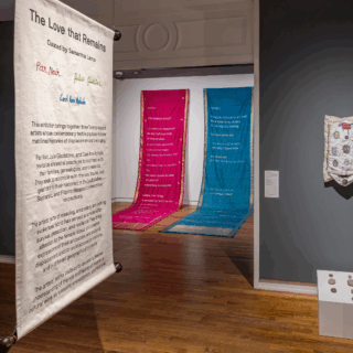Installation view of a text panel in foreground, with pink and blue sarees in the background. To each side are a blue illuminated artwork and a knitted fabric piece.