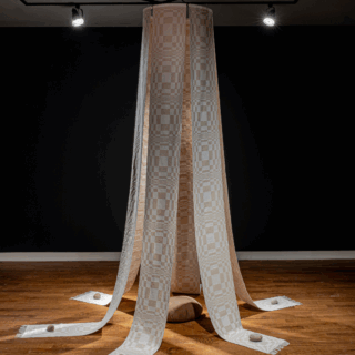 Installation view of long, checkered textile strips hang from the ceiling in a dim gallery. It drapes over a central cushion and spreads outward, anchored by stones.