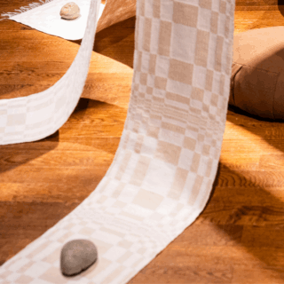Close-up of checkered textile strips cascade onto a wooden floor, anchored by stones. A round cushion sits under where the strips gather.