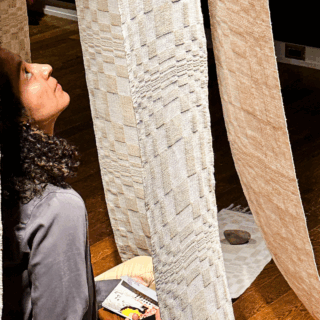 A person sits on a cushion, surrounded by cascading, checkered textile strips. They look up thoughtfully in warm lighting.
