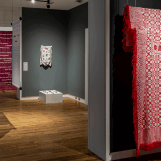 Gallery space with textiles and fabric art on display. Red, blue, and pink pieces with intricate patterns hang against dark walls.