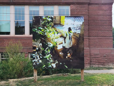 Installation view of Jenelle Smith's photography work "Gia" in front of a red brick wall at 180 Shaw Street. The photograph captures a leafy foreground with a stem that reaches out and covers the face of a person going down the stairs.