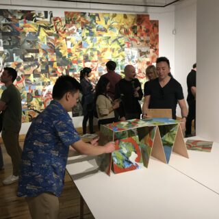 An individual builds a structure using small artworks in a public art program while another person watches, with others viewing artworks displayed on nearby walls.
