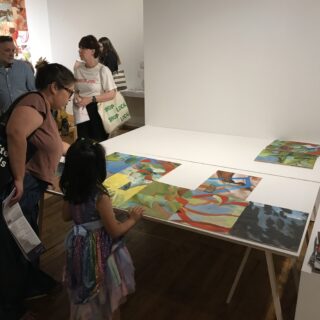 An adult and a young child look at colourful abstract paintings displayed on a white table