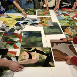 A group of people crowded around a white table arranging small colourful abstract paintings.