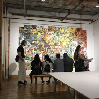 Candid photo of people in a gallery. Four are seated on a bench viewing abstract paintings, one standing nearby, and two others talking while one looks at a piece of paper.