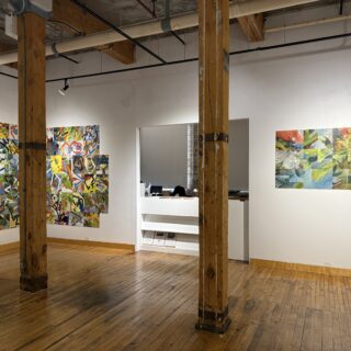 Gallery with wooden floors and exposed beams, showing colourful abstract artworks on white walls and a small desk area in the back.