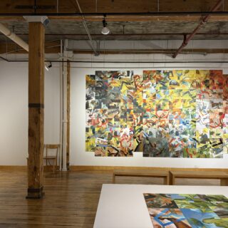 Gallery space with wooden floors and exposed beams, featuring colourful abstract artworks arranged in a grid on a white wall, with benches and a table displaying additional pieces.