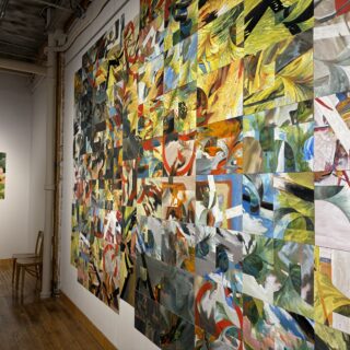 White gallery wall with small colourful abstract paintings arranged in a grid, two wooden chairs sit nearby, with another grid of paintings on an adjacent wall.