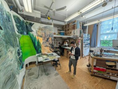 A person is standing in the middle of a studio. In front of hime is a large-scale abstract painting with dominant green colour. The rest of the space is cluttered with art supplies and shelves.