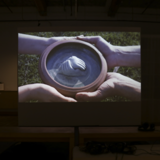 Projected image of two pairs of hands holding a bowl containing a small clay figurine of two hands clasped together, displayed on a large screen in a darkened room.