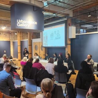 Four panelists sit in chairs while another individual stands with a microphone, addressing the audience. A seated crowd watches the discussion.
