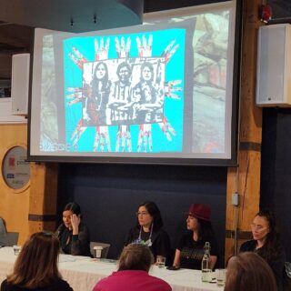 Five individuals sit at a table beneath a projector, speaking to an audience seated in front of them.