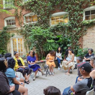 A group of people are all seated outside on chairs, surrounded by trees, all facing and listening attentively to one individual who is speaking.