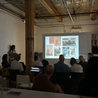 Artist sitting in a gallery speaking to a seated audience. A projection screen beside the artist displays a slide with photographs.