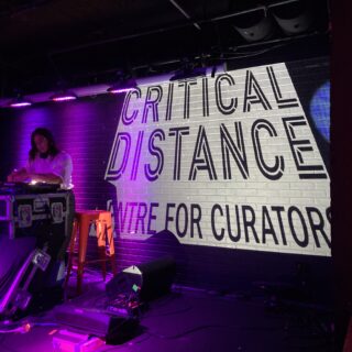 A DJ performs in front of a brick wall illuminated by magenta lighting, with a projected image behind them that reads "Critical Distance Centre for Curators."