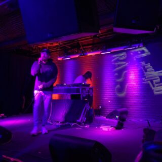 An individual wearing a dark hoodie, glasses, and a baseball cap performs onstage with a microphone. A DJ stands behind them at a turntable setup. Bright lights illuminate the stage.