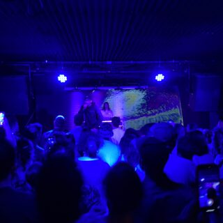 An individual wearing a dark hoodie, glasses, and a baseball cap performs onstage with a microphone. A DJ stands behind them at a turntable setup. Bright stage lights illuminate both the performers and the crowd watching from below.