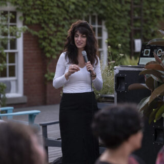 An individual with wavy brown hair, wearing a white long-sleeved V-neck shirt and a black skirt, holds a microphone and addresses a group of people.