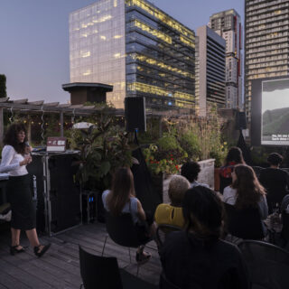 An individual with wavy brown hair, wearing a white long-sleeved V-neck shirt and a black skirt, holds a microphone and addresses a group of people, while a film is being projected onto a screen in front of them.