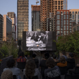 A group of people sit on a rooftop surrounded by plants watching a film projected onto a screen.