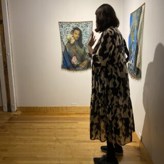 An individual with short, shoulder-length hair, wearing a black and white patterned dress and black sneakers, stands facing an artwork displayed on a white wall.