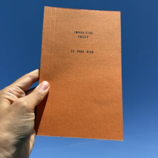 A hand holds an orange book entitled Improvising Freely by Lê Quan Ninh against a bright blue sky.