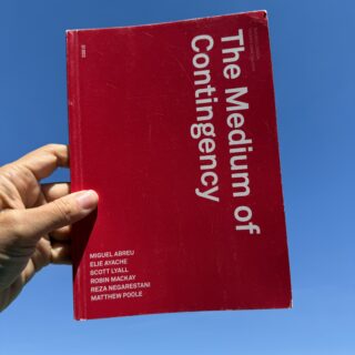 A hand holds a red book titled The Medium of Contingency against a clear blue sky. The cover features large white text, with the names of contributors listed vertically in the lower left corner and gallery details in the upper right.