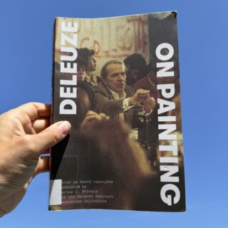 A hand holds a copy of On Painting by Gilles Deleuze against a clear blue sky. The book cover features a blurred image of a man gesturing in a crowd, with the title printed in large white vertical text along both sides of the book.
