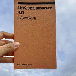 A hand holds an orange book entitled On Contemporary Art by César Aira against a cloudy blue sky. There is a long black horizontal line above the title. The cover also reads the publisher name, "David Zwirner Books," in the bottom left corner.