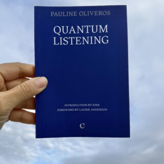 A hand holds a blue book titled Quantum Listening by Pauline Oliveros against a cloudy blue sky. The cover also reads "Introduction by IONE" and "Foreword by Laurie Anderson," with a small circular publisher logo at the bottom.