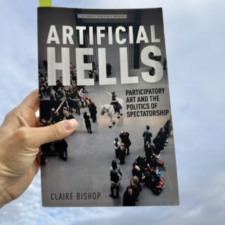 A hand holds a book titled Artificial Hell: Participatory Art and the Politics of Spectatorship. The cover features a photograph of people gathered in a museum-like space, with a green page marker protruding from the top of the book.